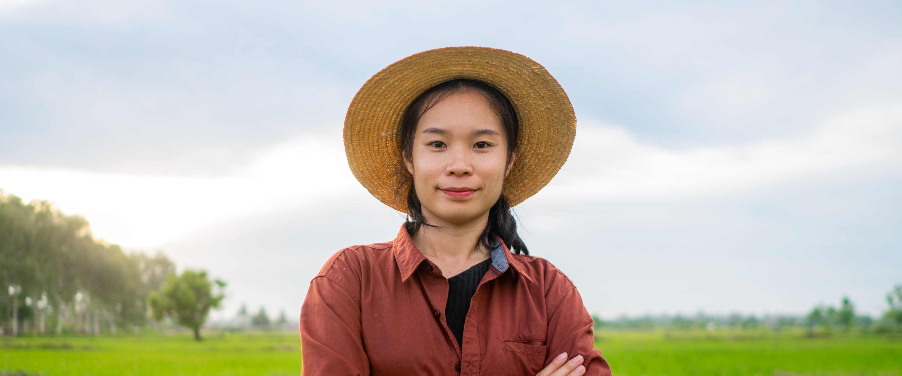 A woman stands confidently with her arms crossed in a green rice field, wearing a wide-brimmed straw hat and a brown shirt. The background features lush greenery and a cloudy sky. The setting conveys a peaceful rural environment.