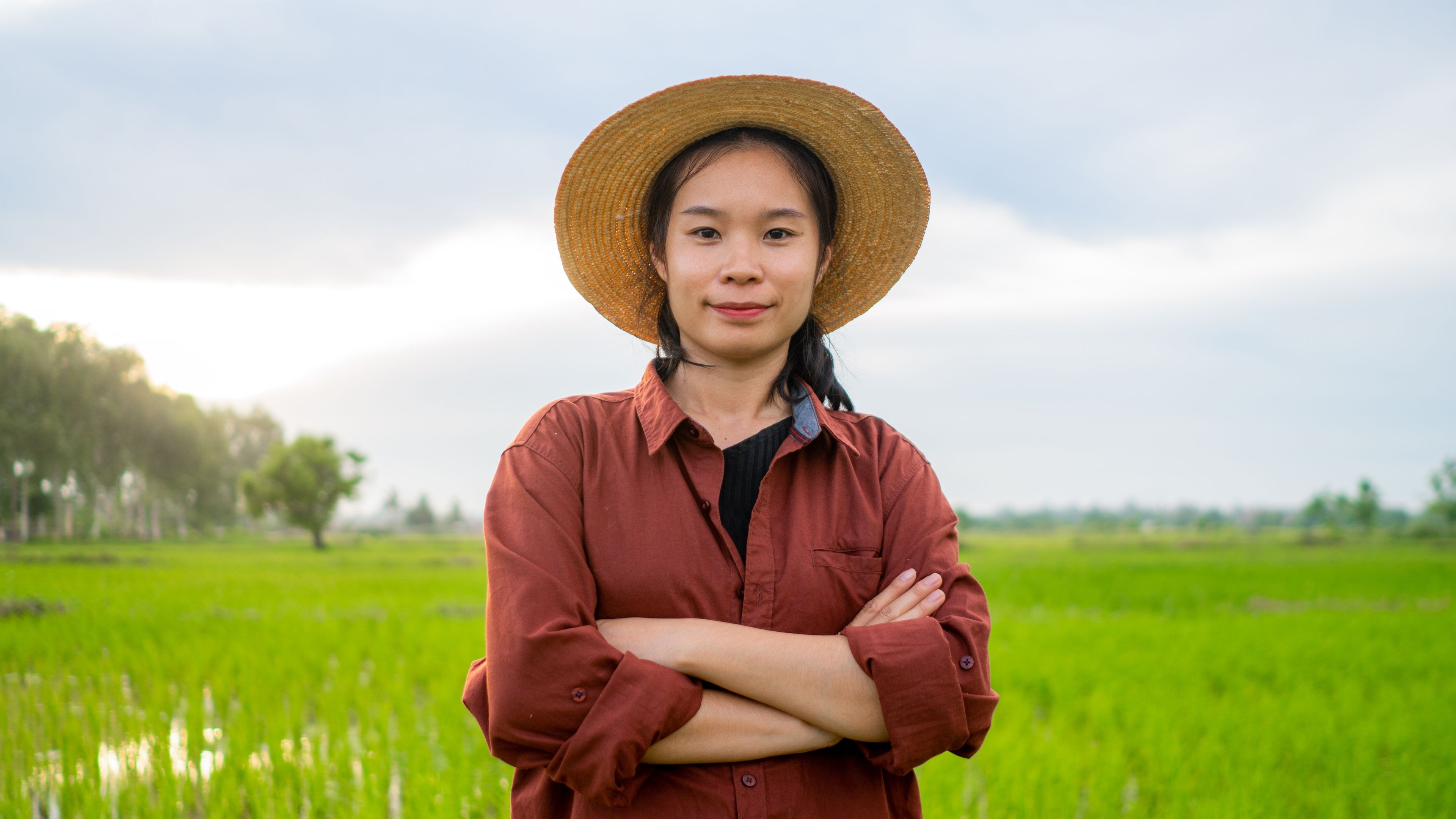 A woman stands confidently with her arms crossed in a green rice field, wearing a wide-brimmed straw hat and a brown shirt. The background features lush greenery and a cloudy sky. The setting conveys a peaceful rural environment.