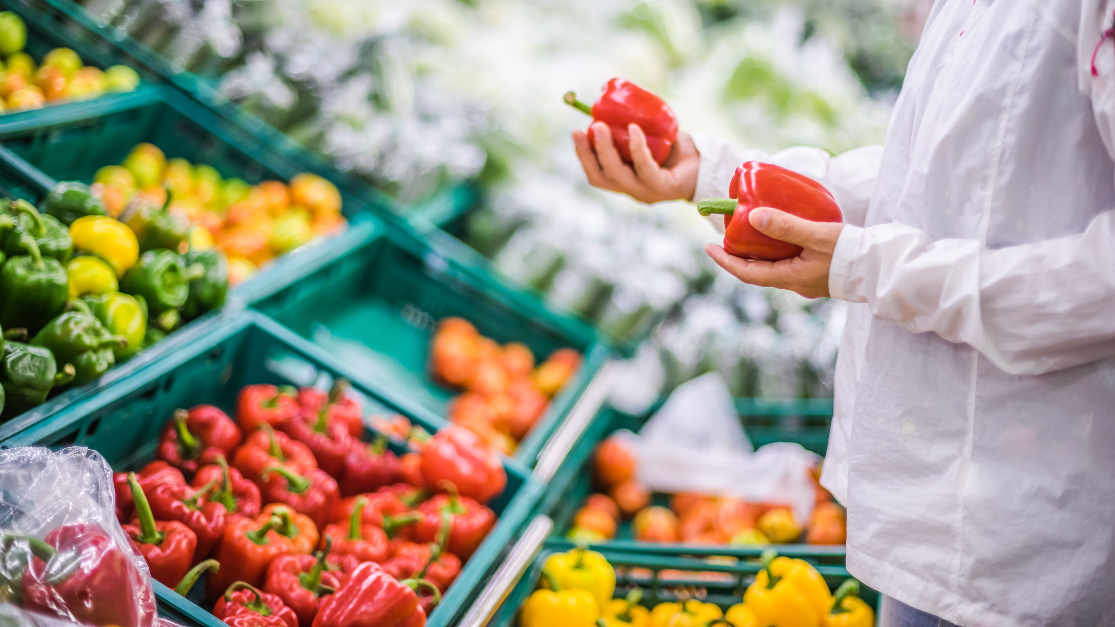 A person wearing a white jacket is selecting red bell peppers from a display in a grocery store. The produce section features various colorful bell peppers, including green and yellow, arranged in green crates. The background shows more fresh produce.