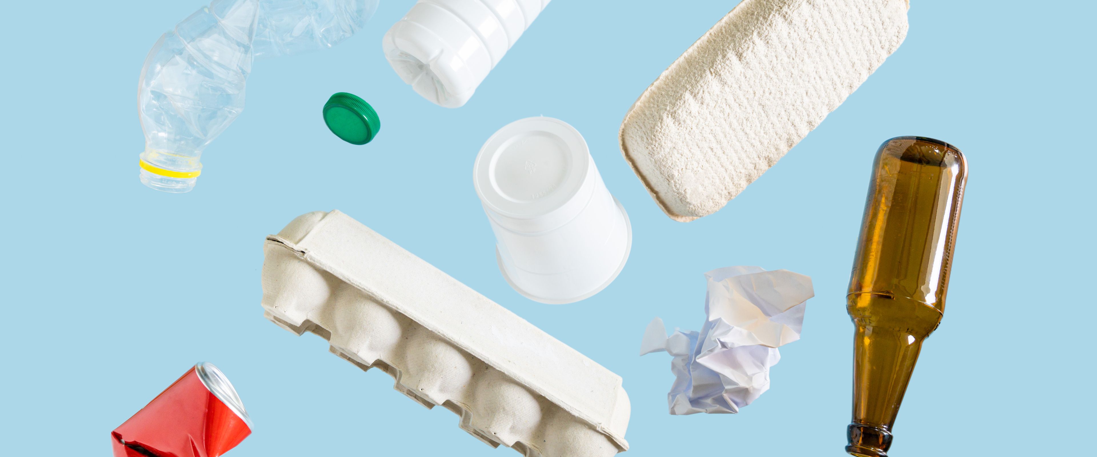 Various recyclable materials, including a plastic bottle, aluminum can, glass bottle, paper egg carton, crumpled paper, and bottle caps, are scattered against a light blue background. The arrangement showcases different types of materials often collected for recycling.