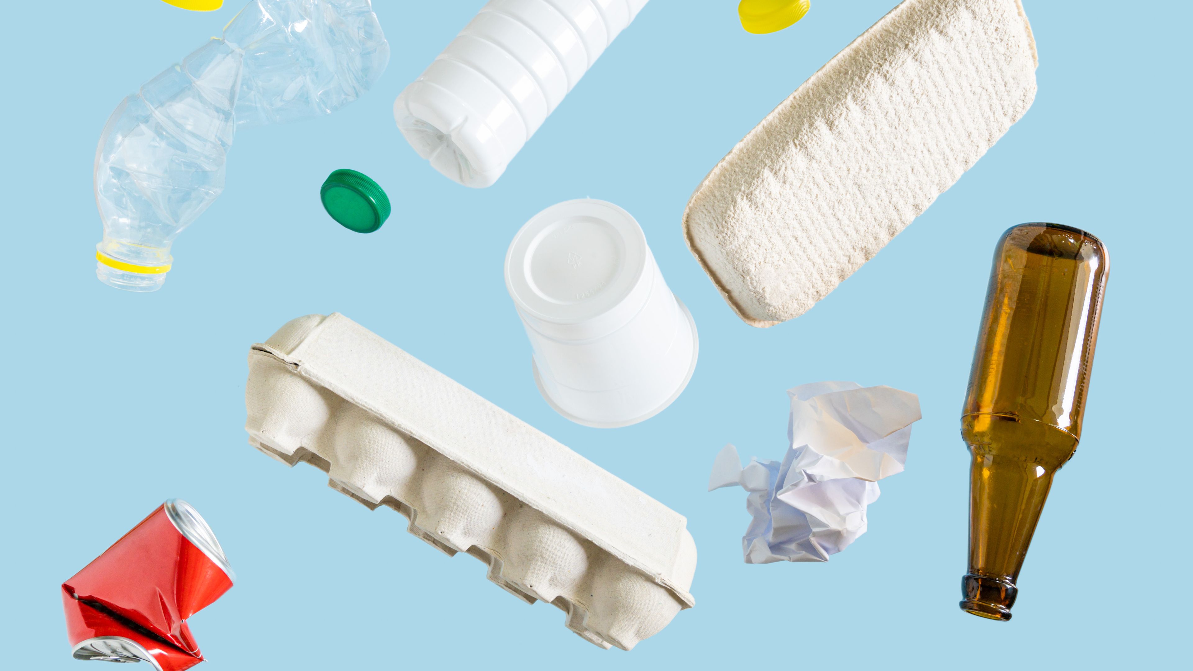 Various recyclable materials, including a plastic bottle, aluminum can, glass bottle, paper egg carton, crumpled paper, and bottle caps, are scattered against a light blue background. The arrangement showcases different types of materials often collected for recycling.