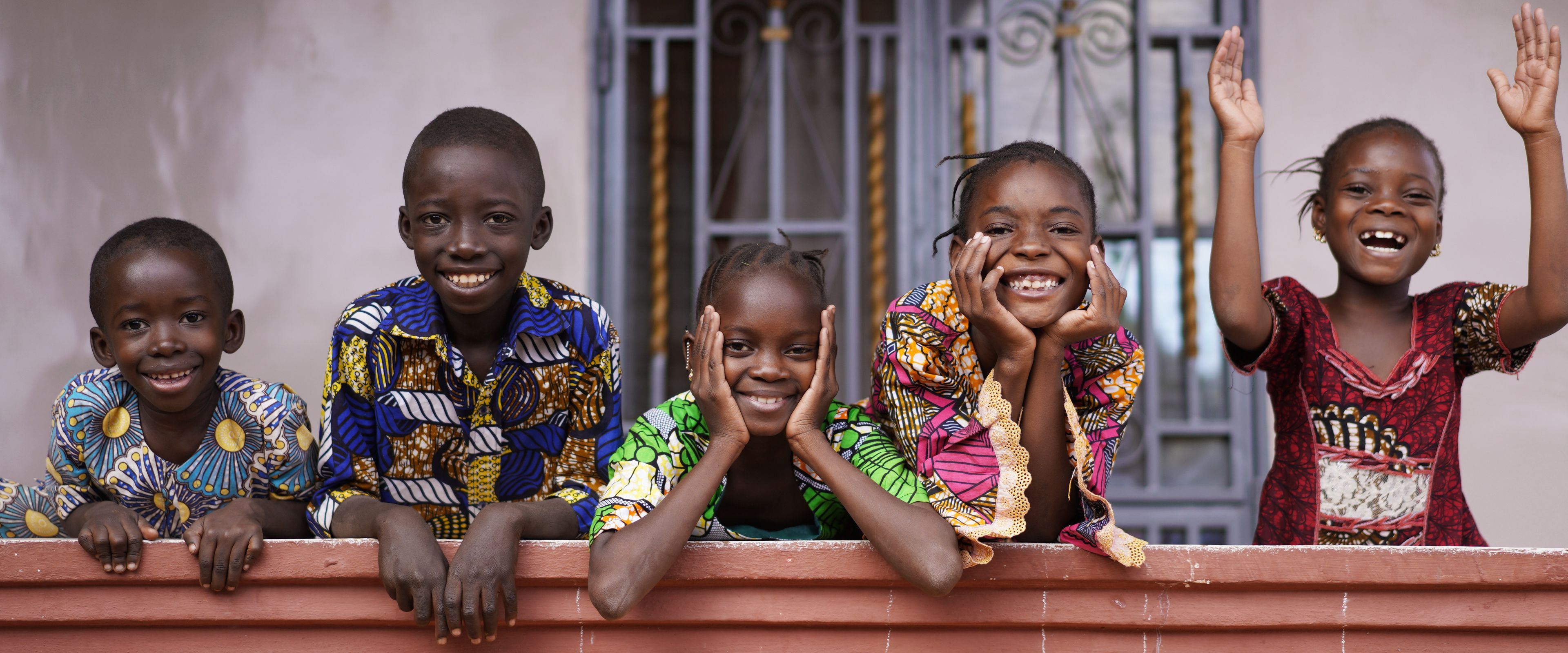 Five children are smiling and leaning on a colorful railing, with a door featuring intricate metal designs in the background. They are dressed in vibrant patterned clothing and radiate joy and energy, with one child raising their hands in excitement.