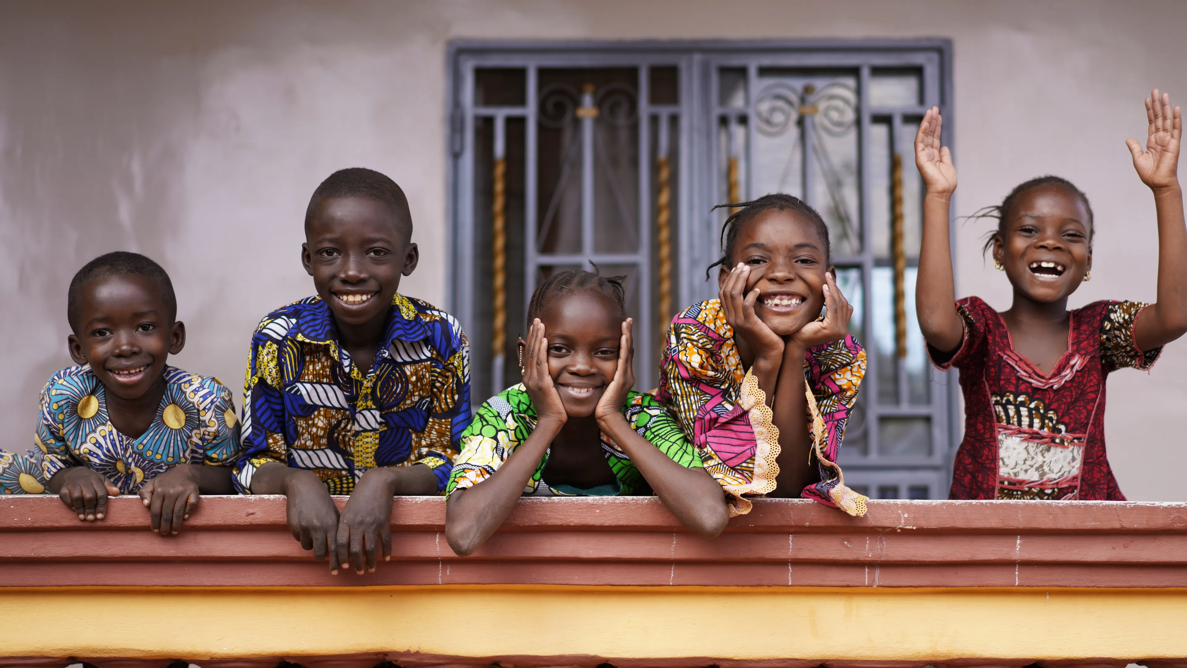 Five children are smiling and leaning on a colorful railing, with a door featuring intricate metal designs in the background. They are dressed in vibrant patterned clothing and radiate joy and energy, with one child raising their hands in excitement.