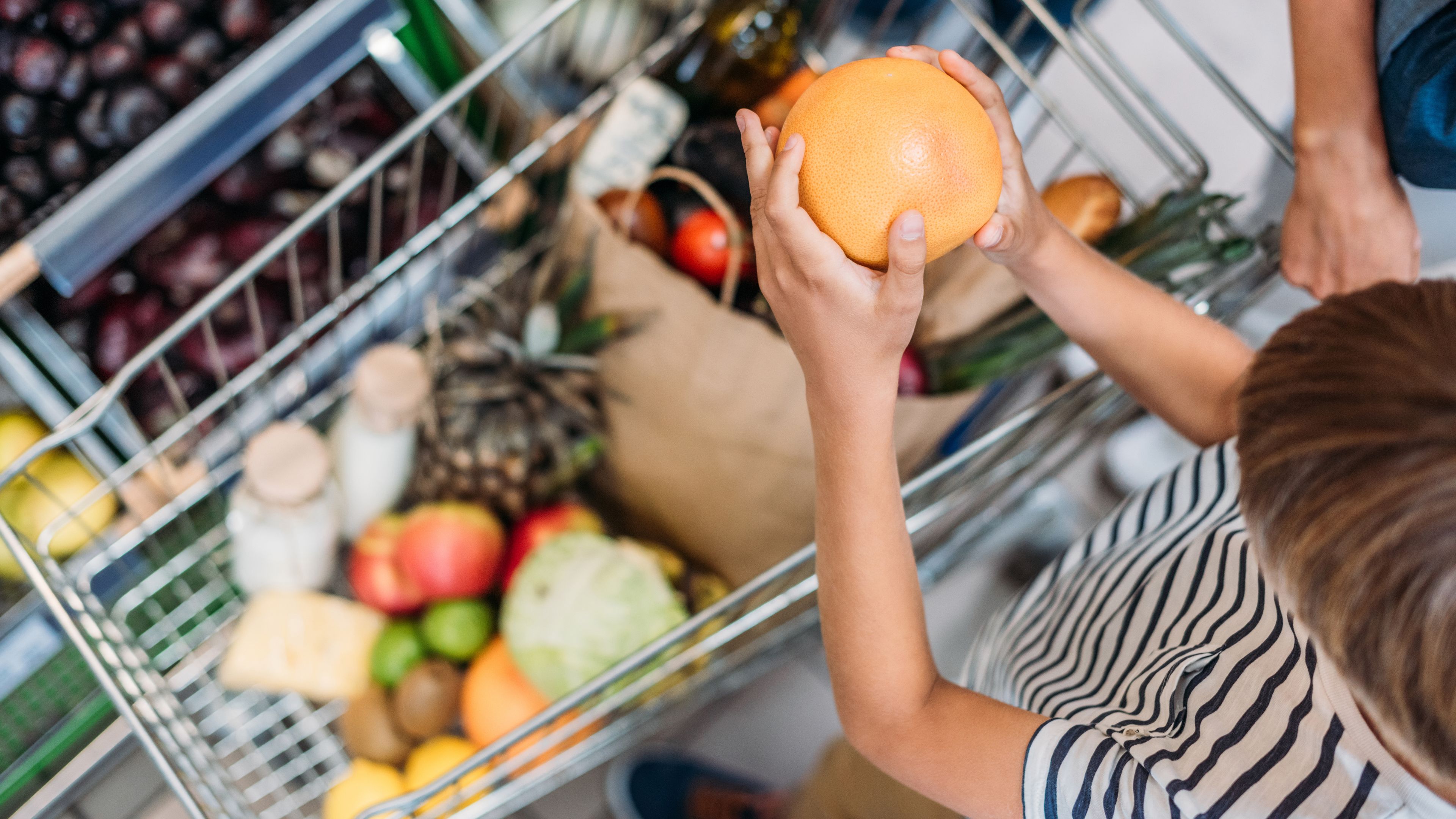 A child holding an orange stands next to a shopping cart filled with various groceries, including fruits, vegetables, and bottles. The cart is packed with colorful and fresh produce. Another person's hand is partially visible, assisting.