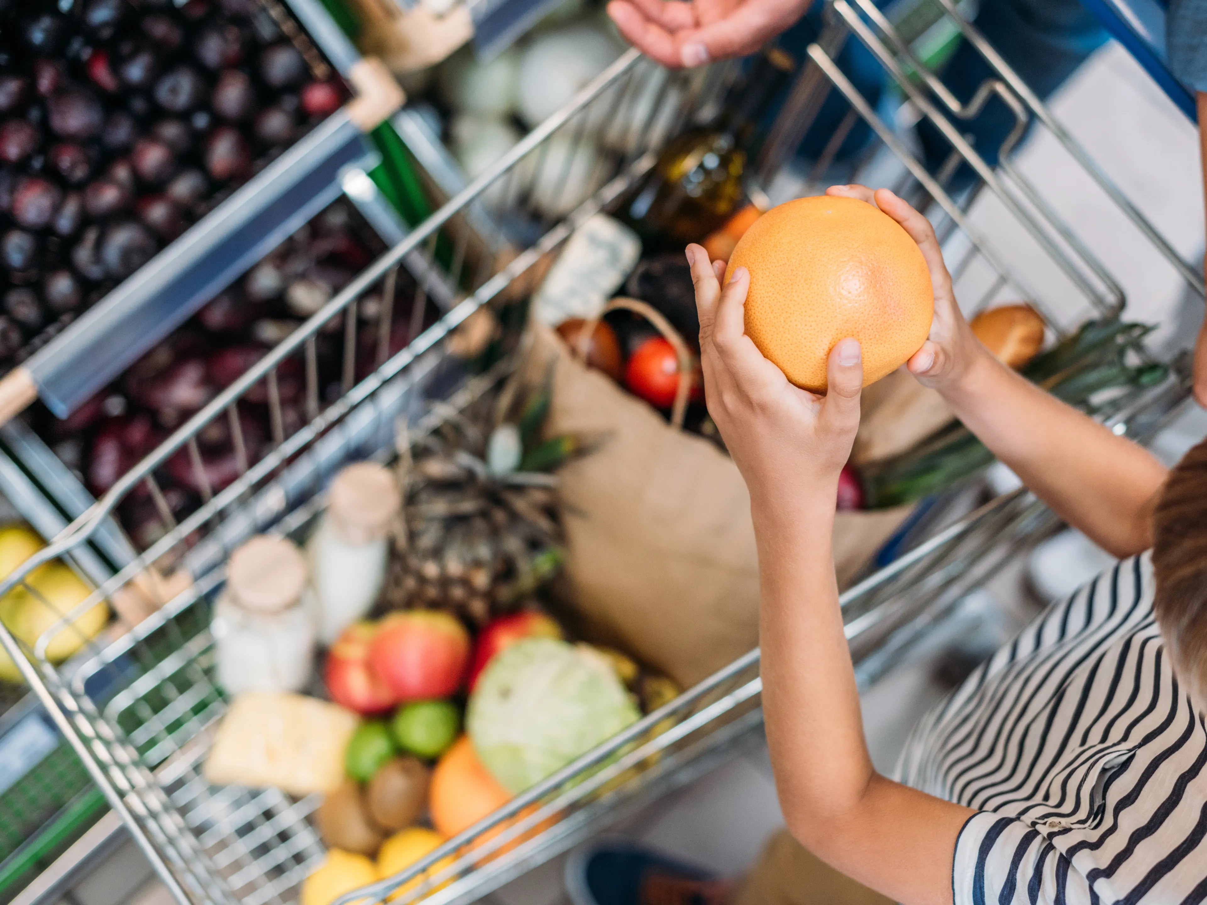 A child holding an orange stands next to a shopping cart filled with various groceries, including fruits, vegetables, and bottles. The cart is packed with colorful and fresh produce. Another person's hand is partially visible, assisting.