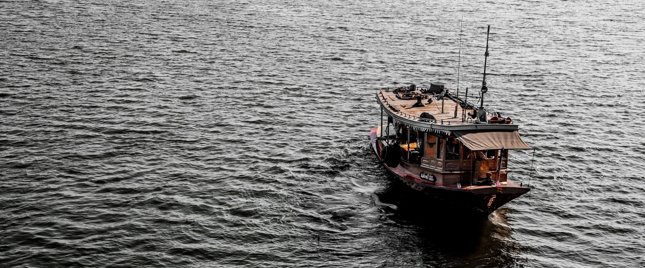 A wooden boat navigates calm gray waters, creating gentle ripples as it moves. The boat has a cabin structure and is equipped with a mast and ropes. The surrounding scene features expansive water with no visible land or sky elements.