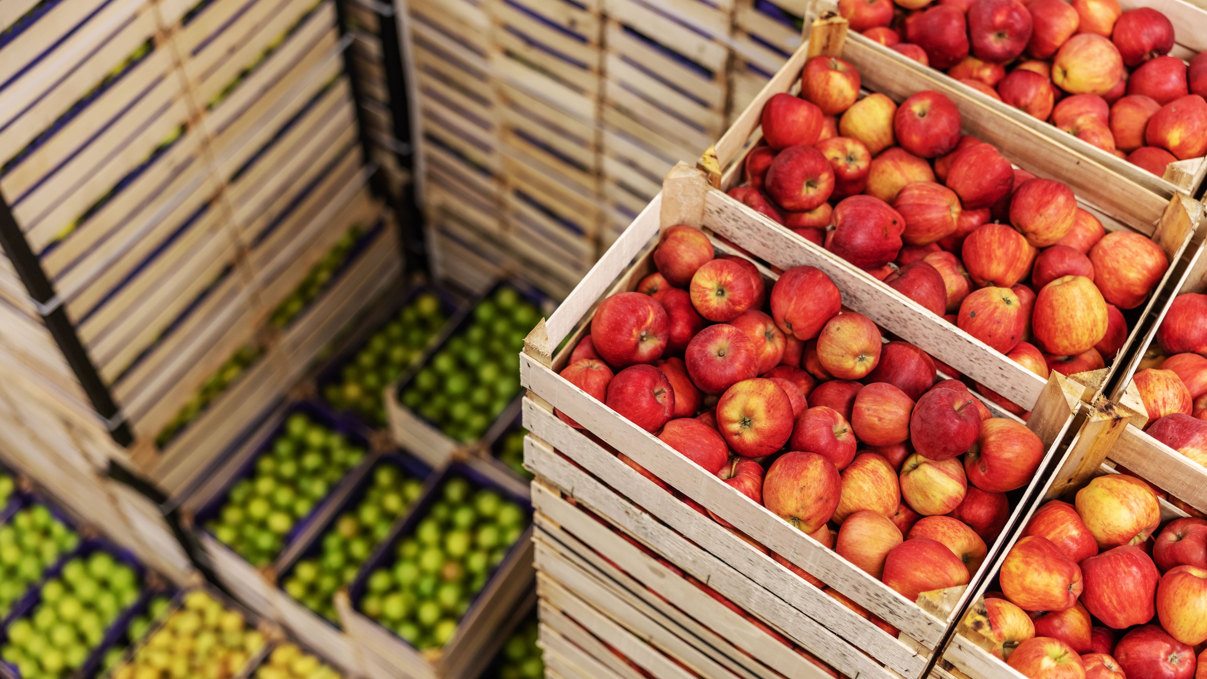 Stacks of wooden crates filled with red apples are shown, alongside crates containing green apples and other fruits. The crates are neatly arranged in a warehouse setting. The view is angled from above, emphasizing the organized layout.