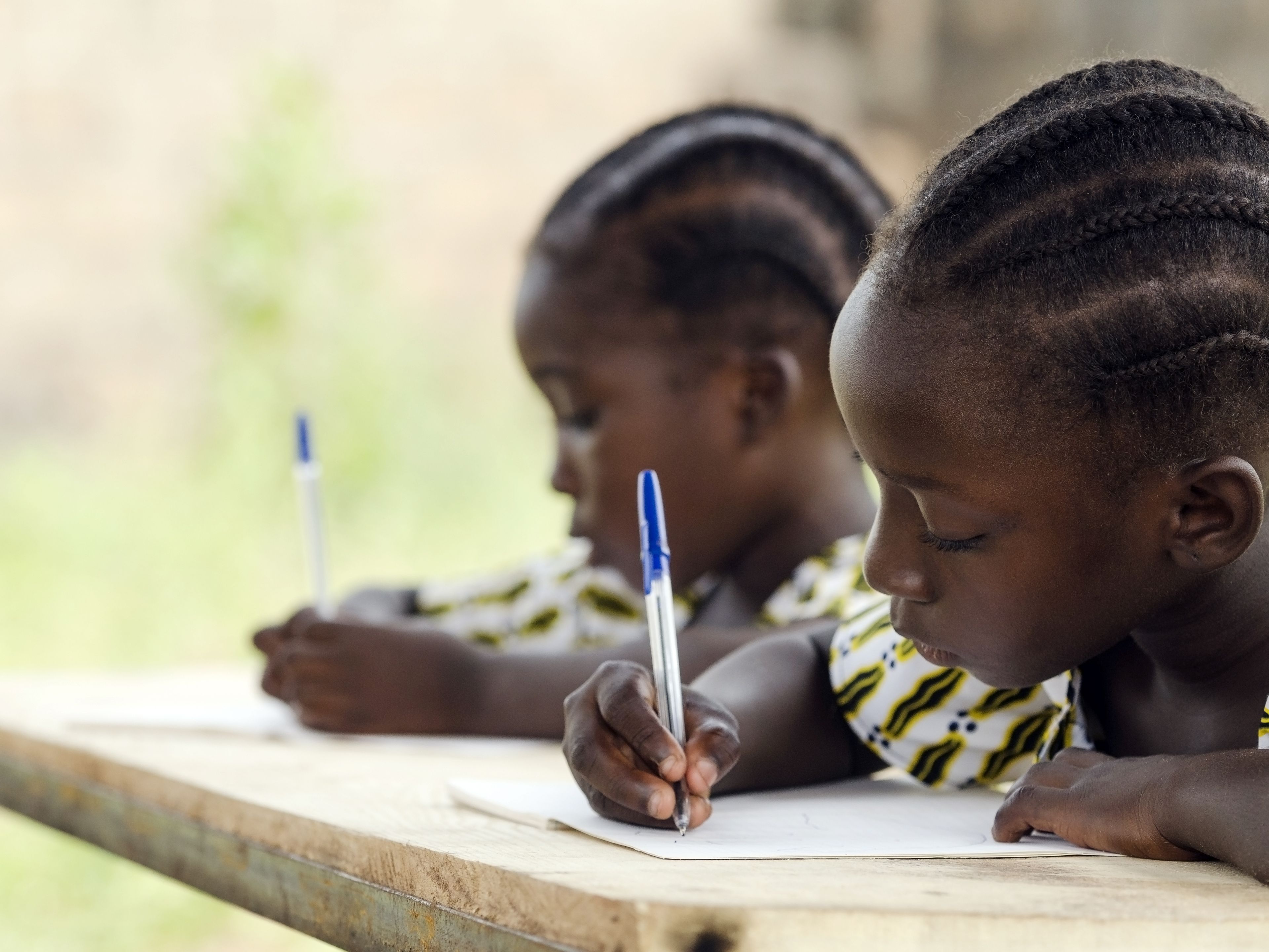 Two young girls are seated at a wooden desk outdoors, focused on writing in notebooks with pens. They are dressed in patterned, matching outfits. The background is blurred, highlighting the natural setting.