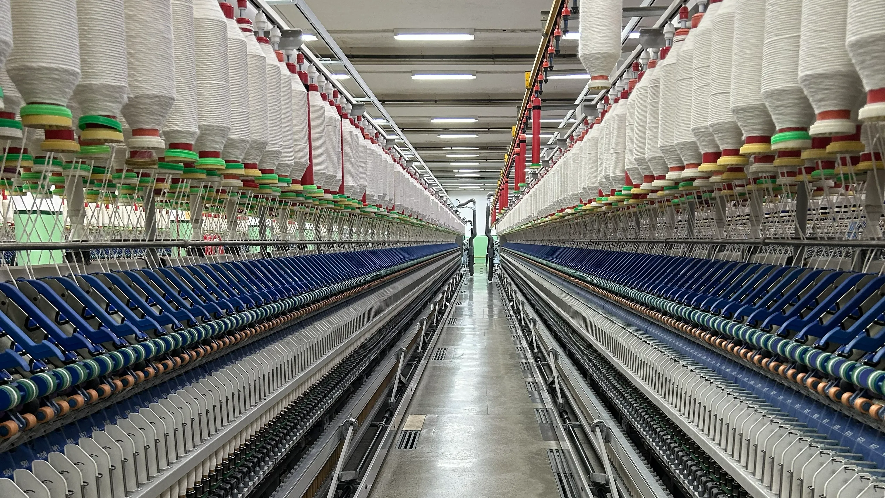 A long perspective view of a textile factory production line with spinning machinery lining both sides. Spools of thread in white and various colors are mounted on top, while the machinery processes the threads below. The floor is shiny and clean, and the space is brightly lit with fluorescent lights.