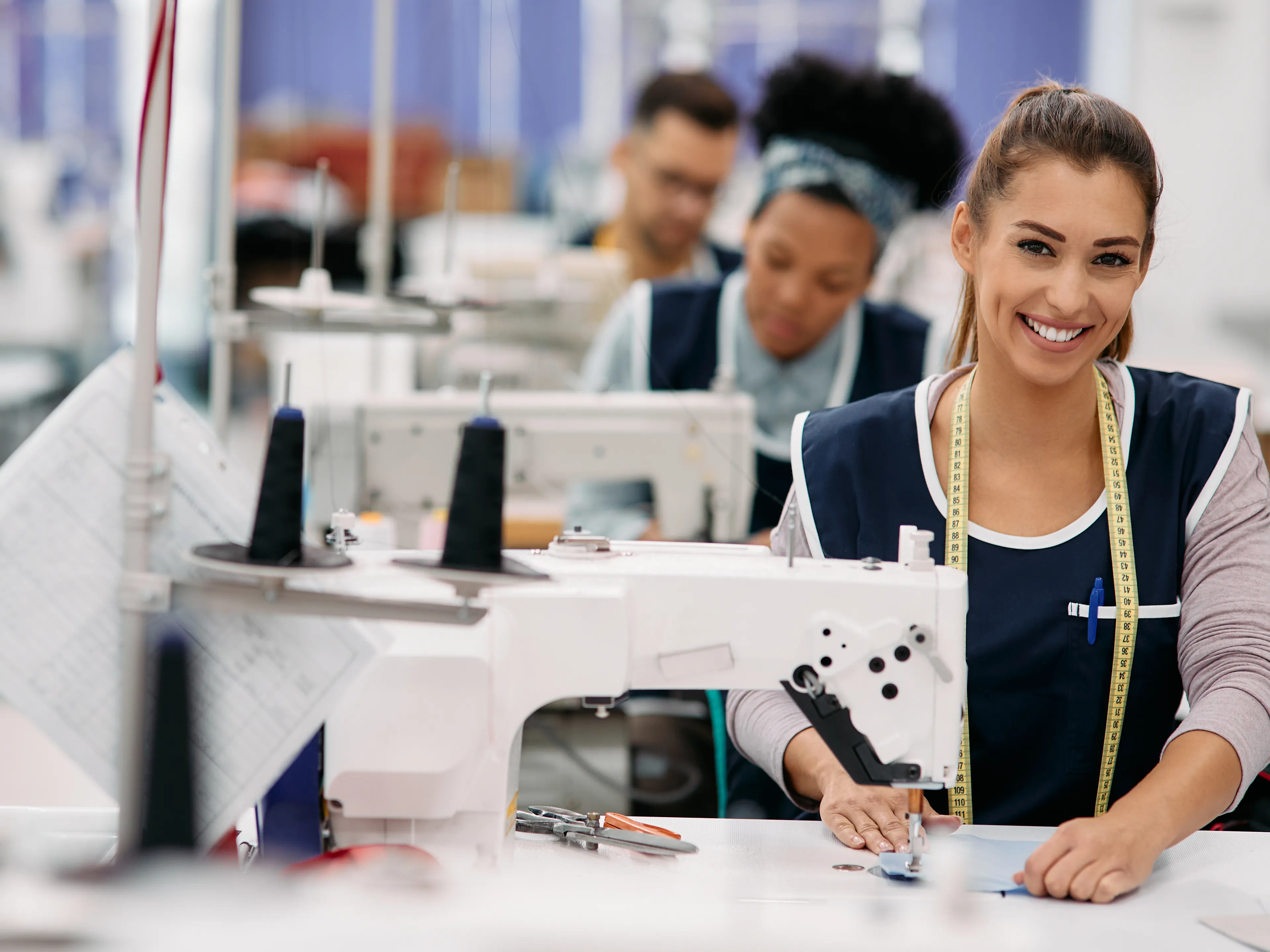 A smiling woman works at a sewing machine in a textile factory, wearing a tape measure around her neck. She is joined by colleagues in the background who are also focused on their tasks. The workspace is bright and organized, with tools and materials visible on the table.