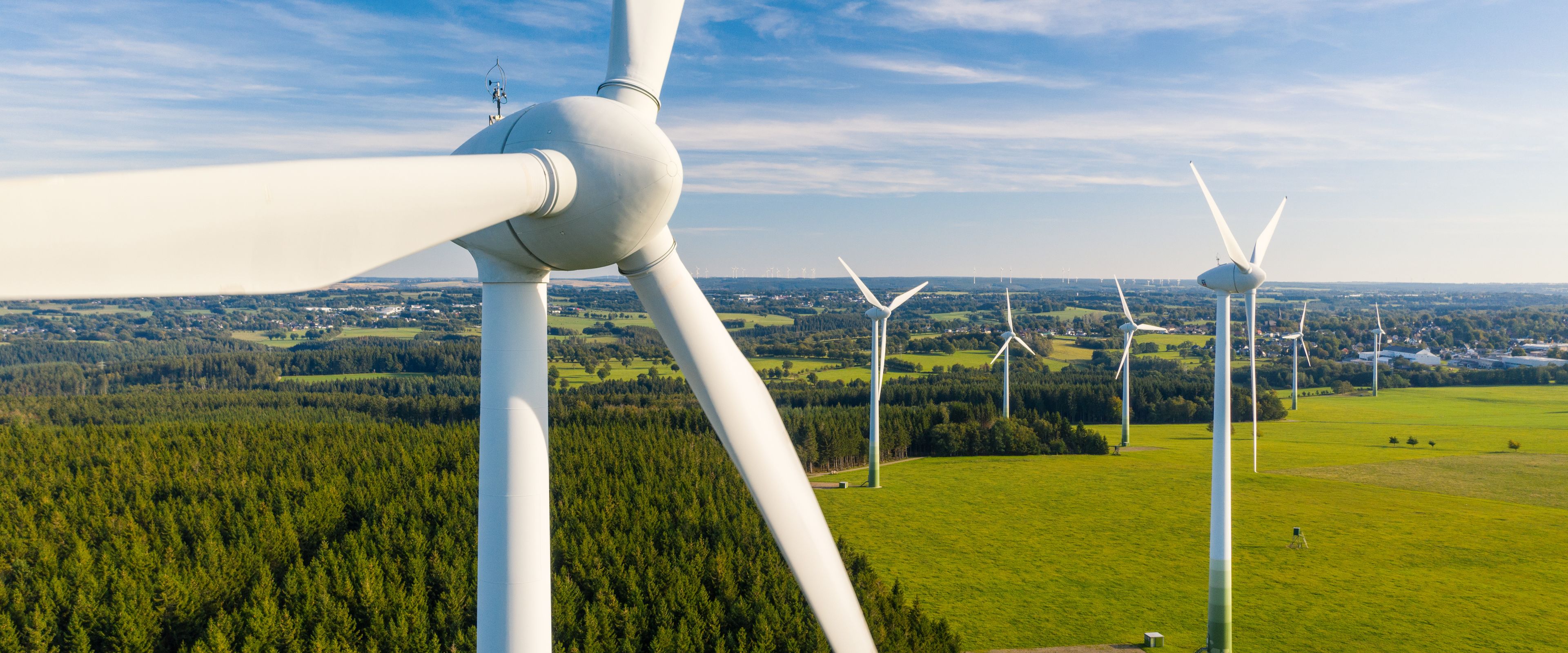 A scenic view of a wind farm with multiple white wind turbines standing tall in a grassy field surrounded by dense forests. The sky is clear and bright blue with scattered clouds. The landscape stretches into the distance, showcasing green fields and distant trees.