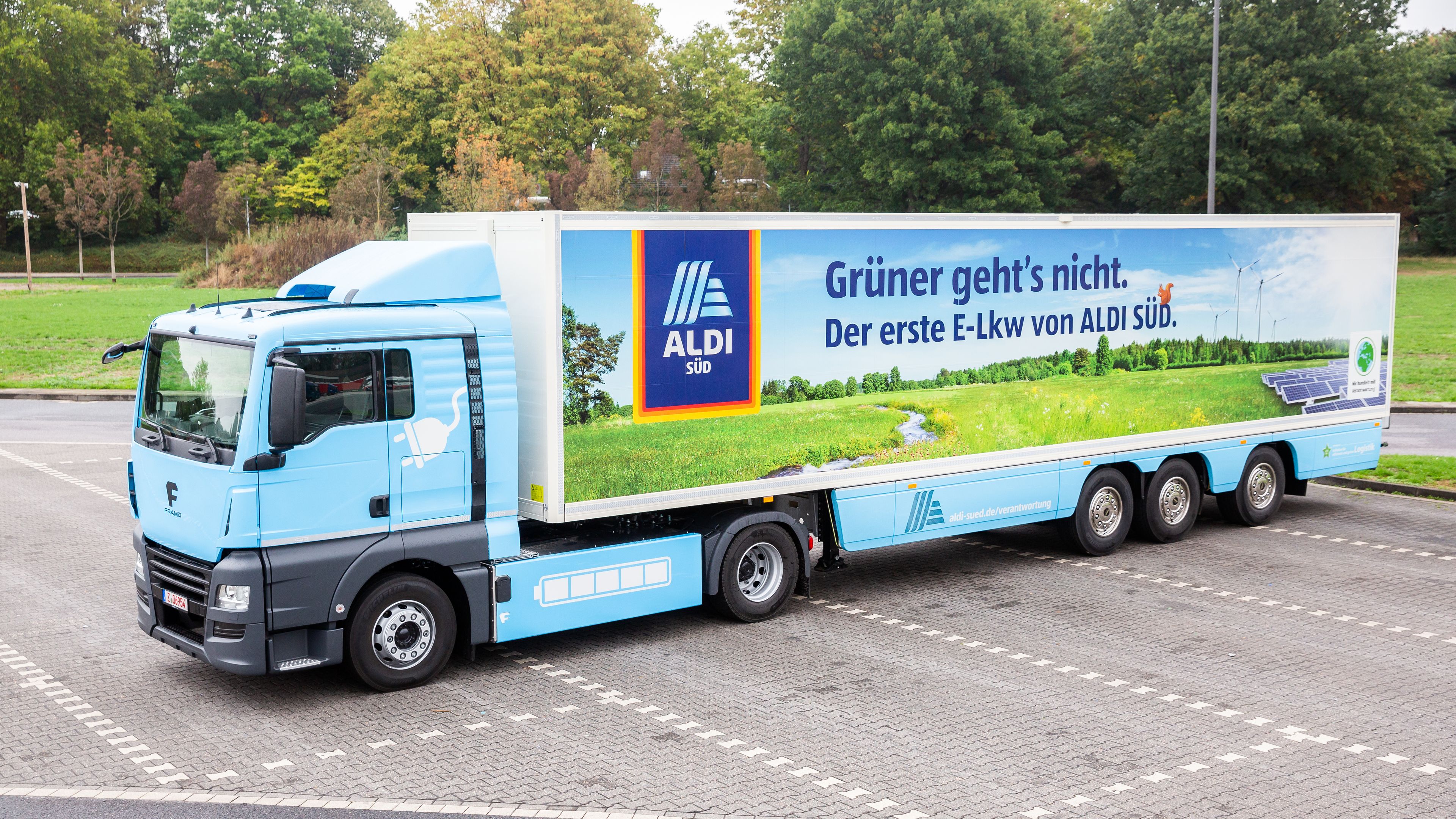 A large blue and white electric truck is parked in a lot, bearing the ALDI Süd logo and the message "Grüner geht's nicht. Der erste E-Lkw von ALDI Süd" promoting sustainability. Green grass and trees are visible in the background, emphasizing eco-friendly themes.