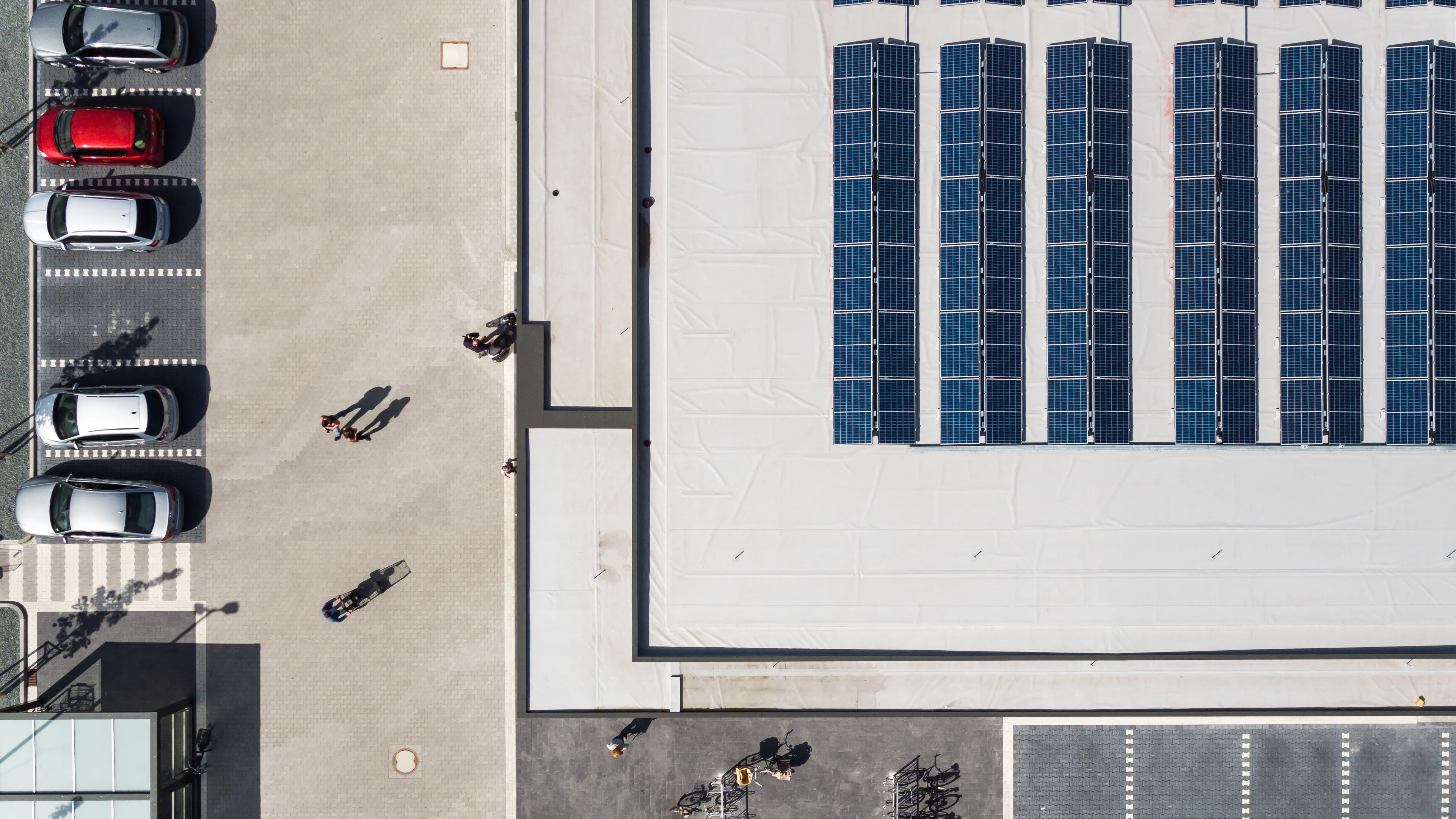 A top-down view of a modern building with solar panels installed on its roof. To the left, there is a parking lot with several parked cars, and people walking on the paved area between the building and the parking lot. The scene is well-lit, indicating a sunny day.