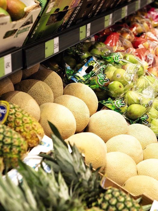 A well-organized grocery store produce section displaying a variety of fruits. Pineapples, melons, pears, and apples are neatly arranged, some wrapped in plastic packaging. The aisle is brightly lit, showcasing the fresh and colorful selection.