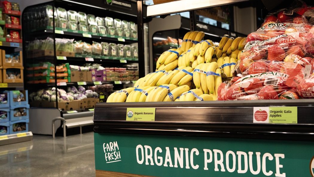 A supermarket produce section featuring a display of organic bananas and bags of organic Gala apples. Signs indicating "Farm Fresh" and "Organic Produce" are visible. Shelves in the background hold an assortment of other fresh organic vegetables and products.