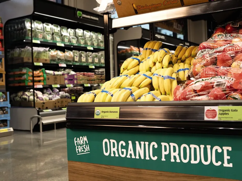 A supermarket produce section featuring a display of organic bananas and bags of organic Gala apples. Signs indicating "Farm Fresh" and "Organic Produce" are visible. Shelves in the background hold an assortment of other fresh organic vegetables and products.