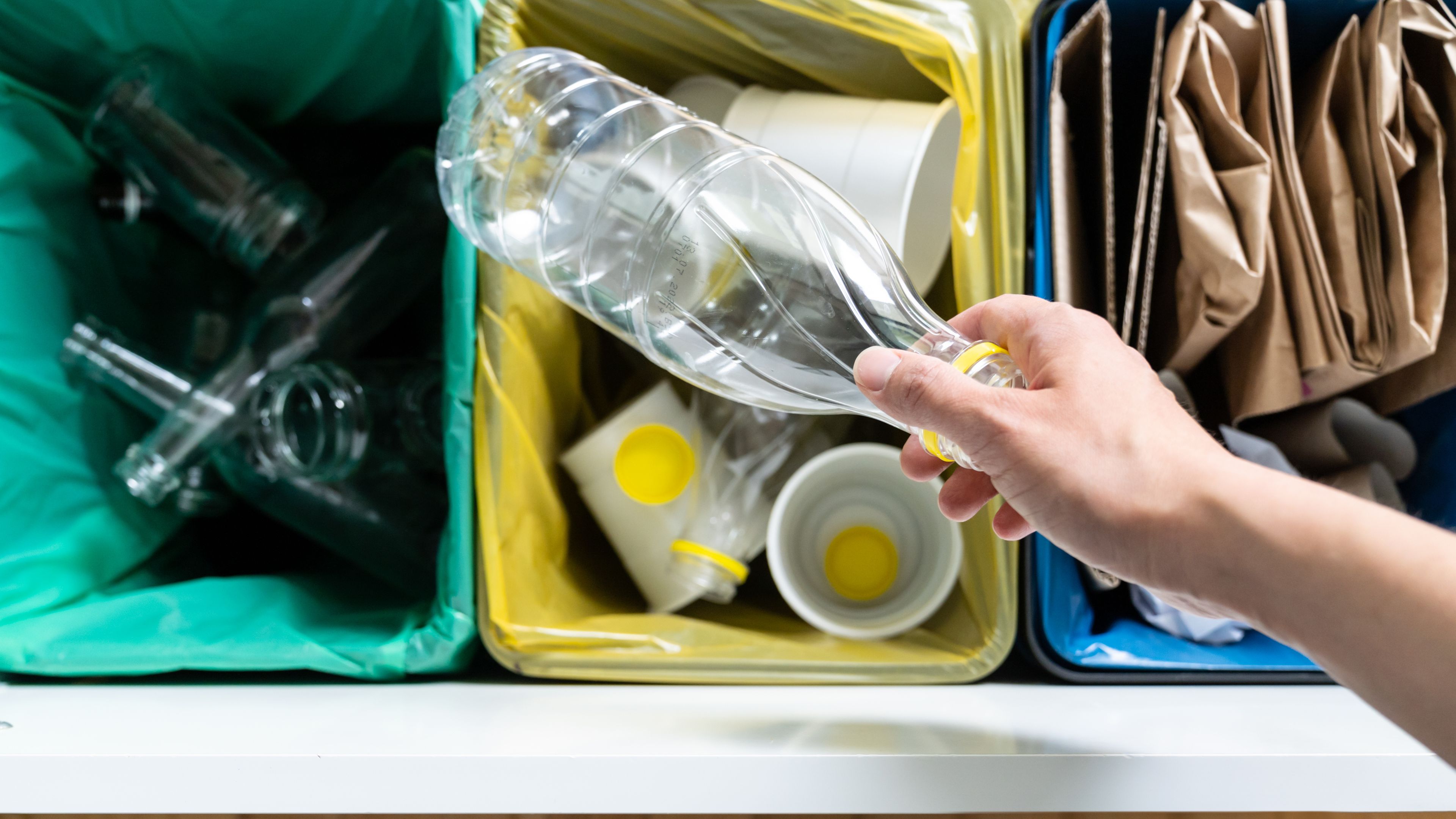 A person's hand holding a plastic bottle over a set of three recycling bins. The bins are color-coded and contain glass, plastic, and paper items. The scene promotes waste sorting and recycling.