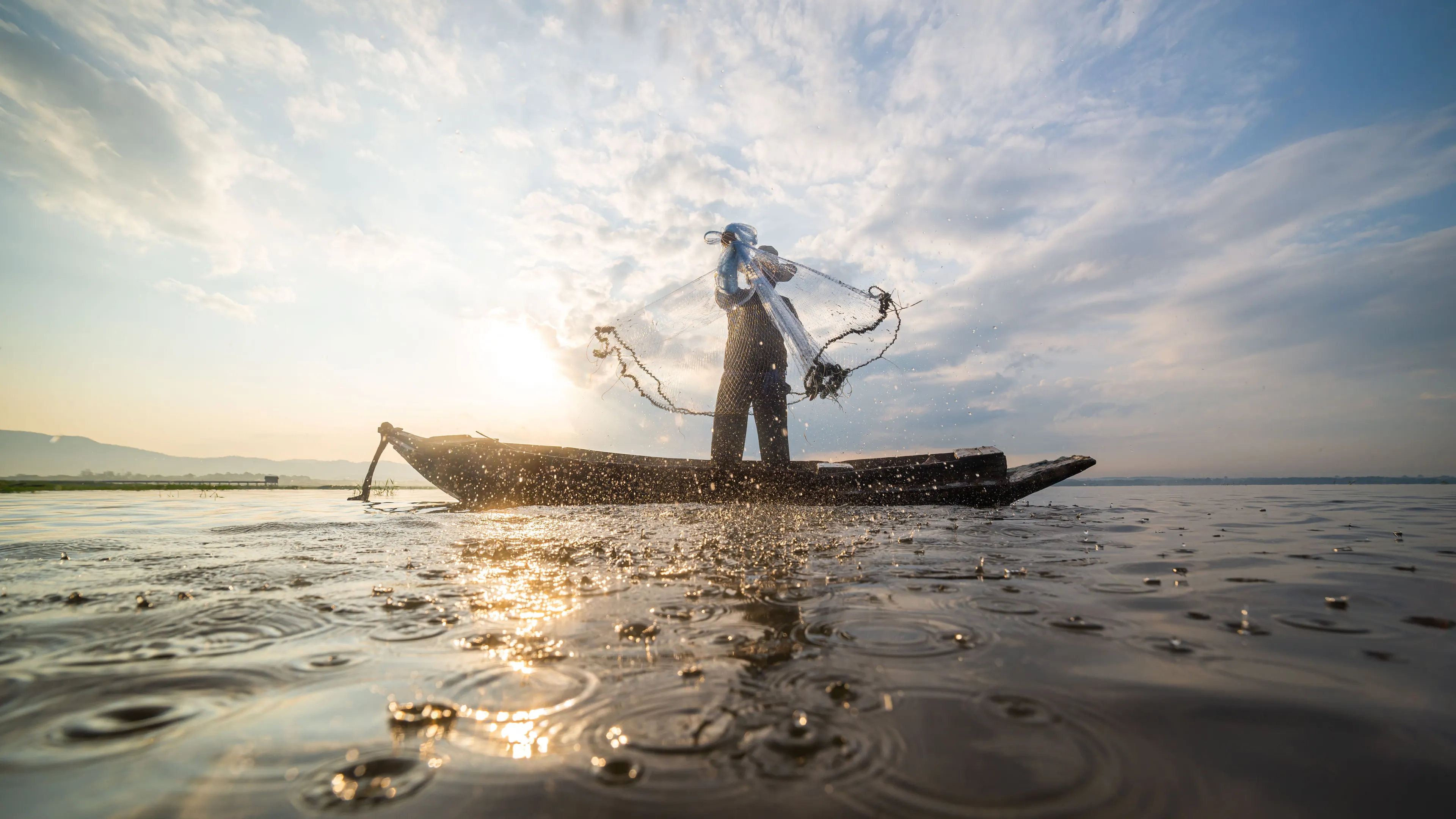 A fisherman standing on a small wooden boat throws a fishing net into the water under a dramatic, cloudy sky at sunrise or sunset. The water reflects the golden light, and droplets splash as the net spreads out. The tranquil scene conveys a sense of peaceful labor.