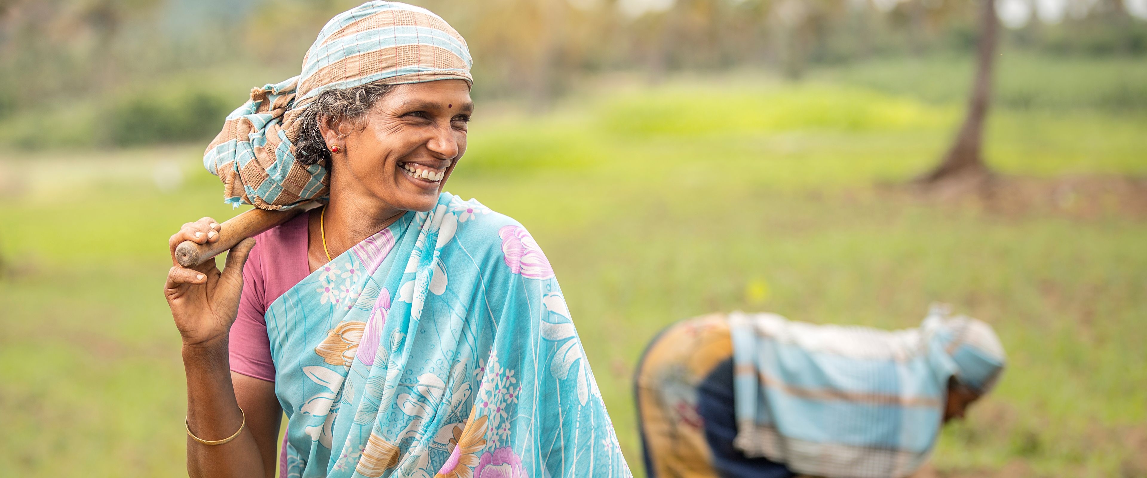 A woman wearing a colorful saree and headscarf smiles while holding a farm tool over her shoulder in a rural field. Another person in the background is bent over, tending to the soil. The scene depicts agricultural work under bright, natural lighting.