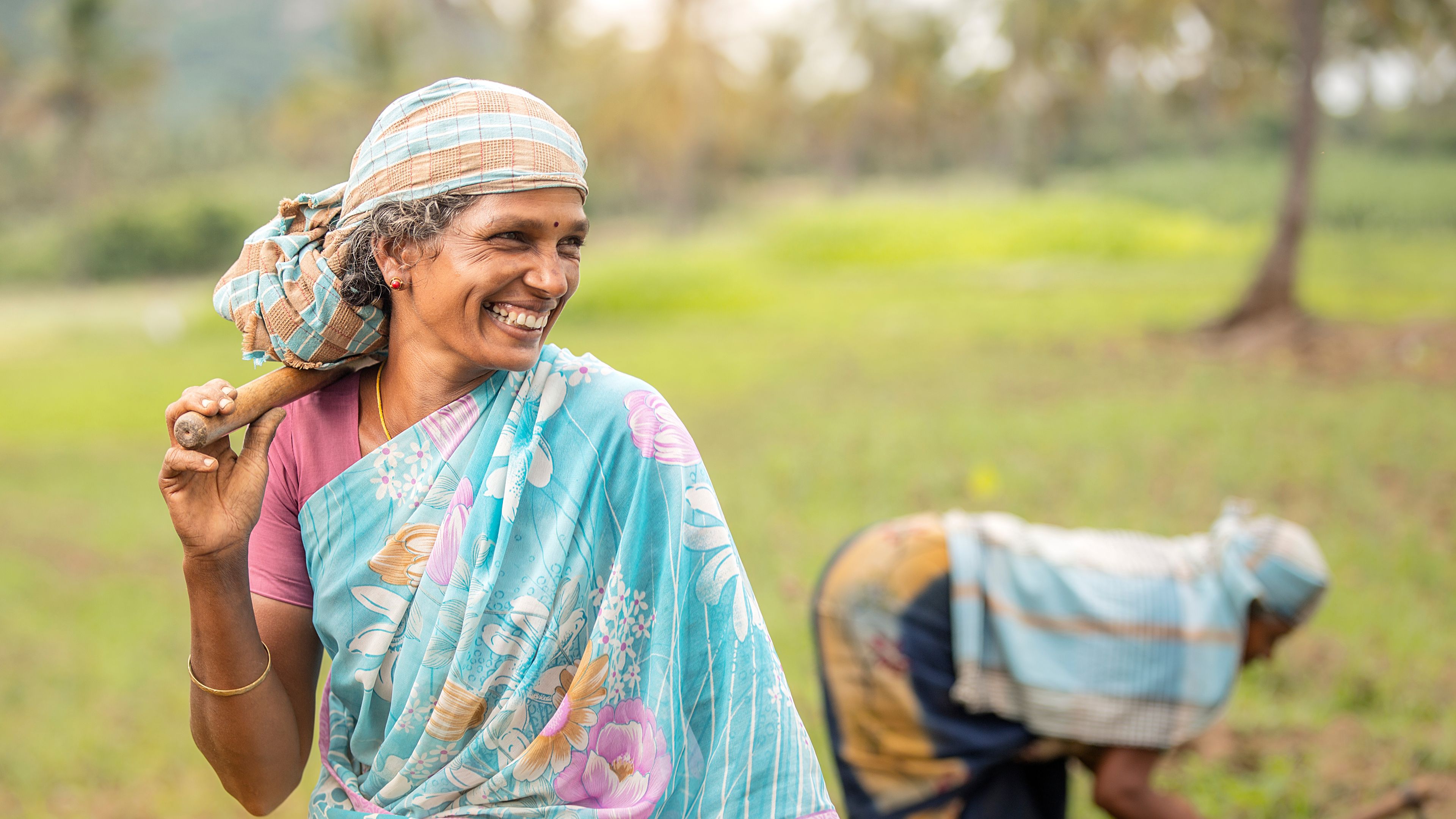 A woman wearing a colorful saree and headscarf smiles while holding a farm tool over her shoulder in a rural field. Another person in the background is bent over, tending to the soil. The scene depicts agricultural work under bright, natural lighting.