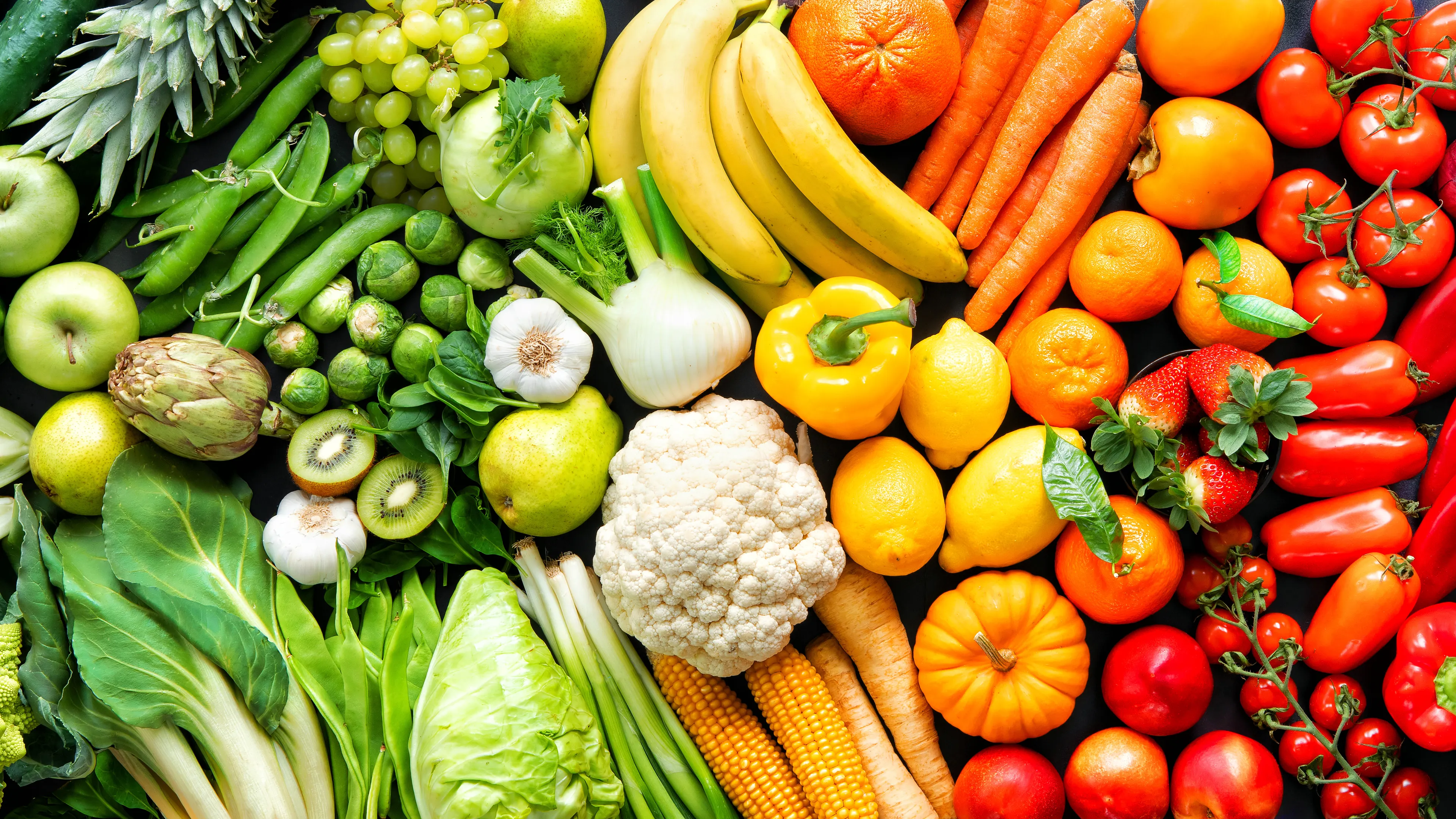 An array of fresh fruits and vegetables arranged in a rainbow gradient, starting with green leafy produce on the left and transitioning through yellow, orange, red, and purple items to the right. It includes lettuce, bananas, oranges, peppers, eggplants, and grapes. The vibrant colors emphasize the diversity of fresh, healthy produce.