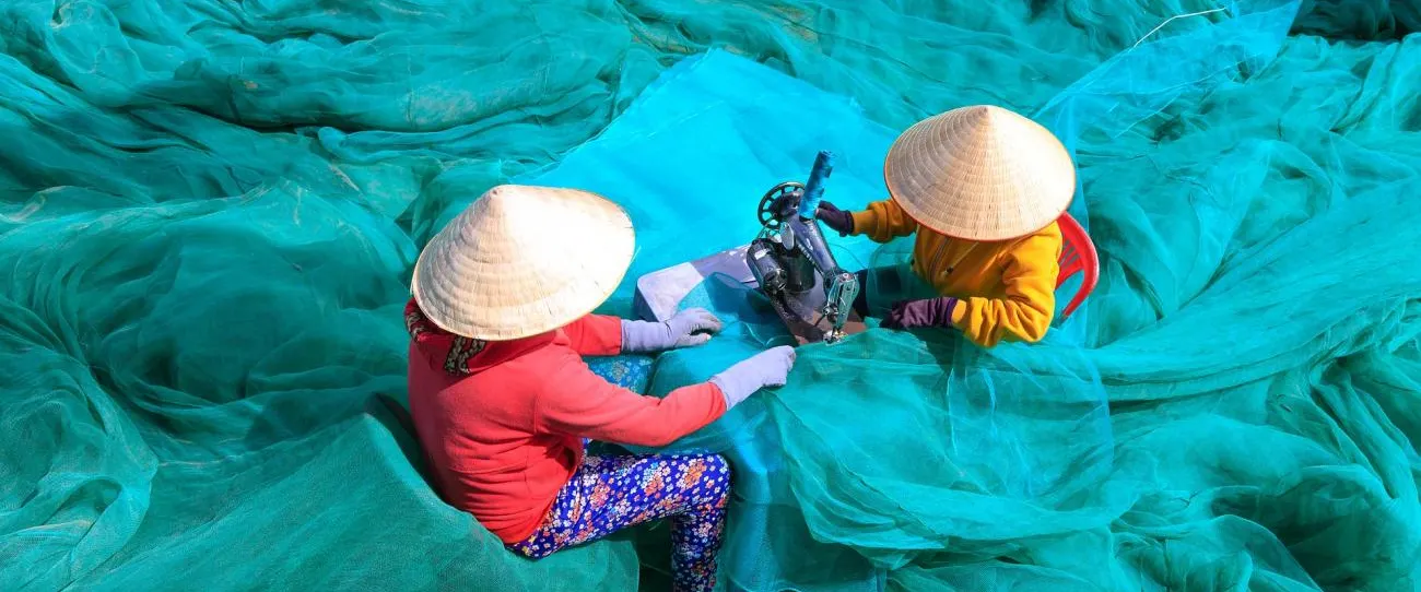 Two people wearing conical hats are sitting amidst a large expanse of bright green fishing nets. They are working together with a sewing machine to repair the nets. The scene highlights labor and craftsmanship in a vibrant outdoor setting.
