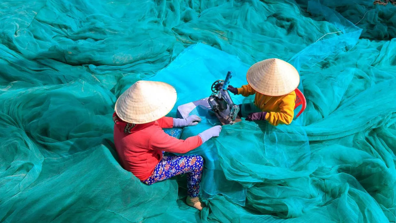 Two people wearing conical hats are sitting amidst a large expanse of bright green fishing nets. They are working together with a sewing machine to repair the nets. The scene highlights labor and craftsmanship in a vibrant outdoor setting.