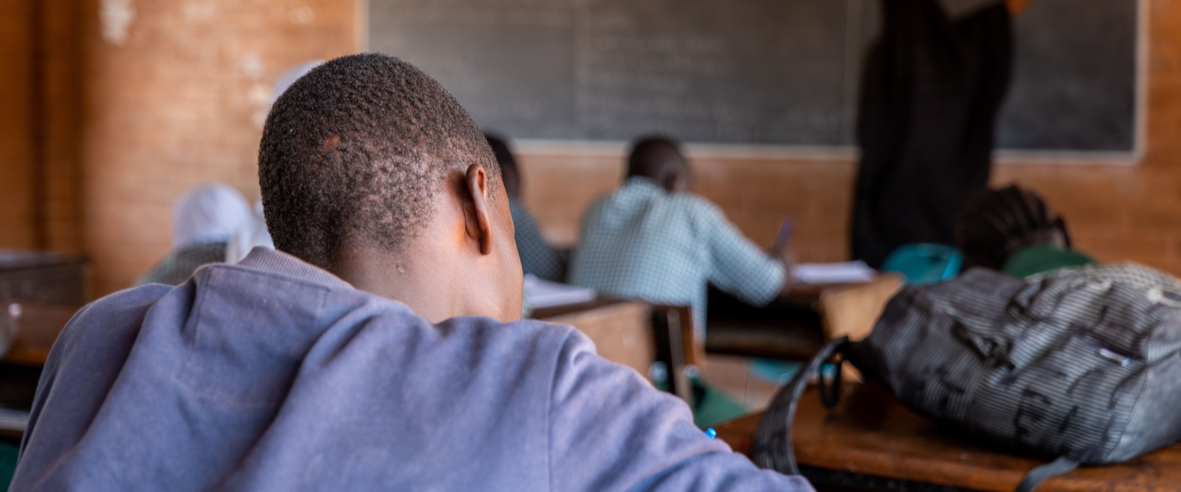 A classroom scene with students seated at desks, focusing on a blackboard at the front. A teacher, seen from behind, writes on the board. Backpacks and learning materials are visible on the desks.