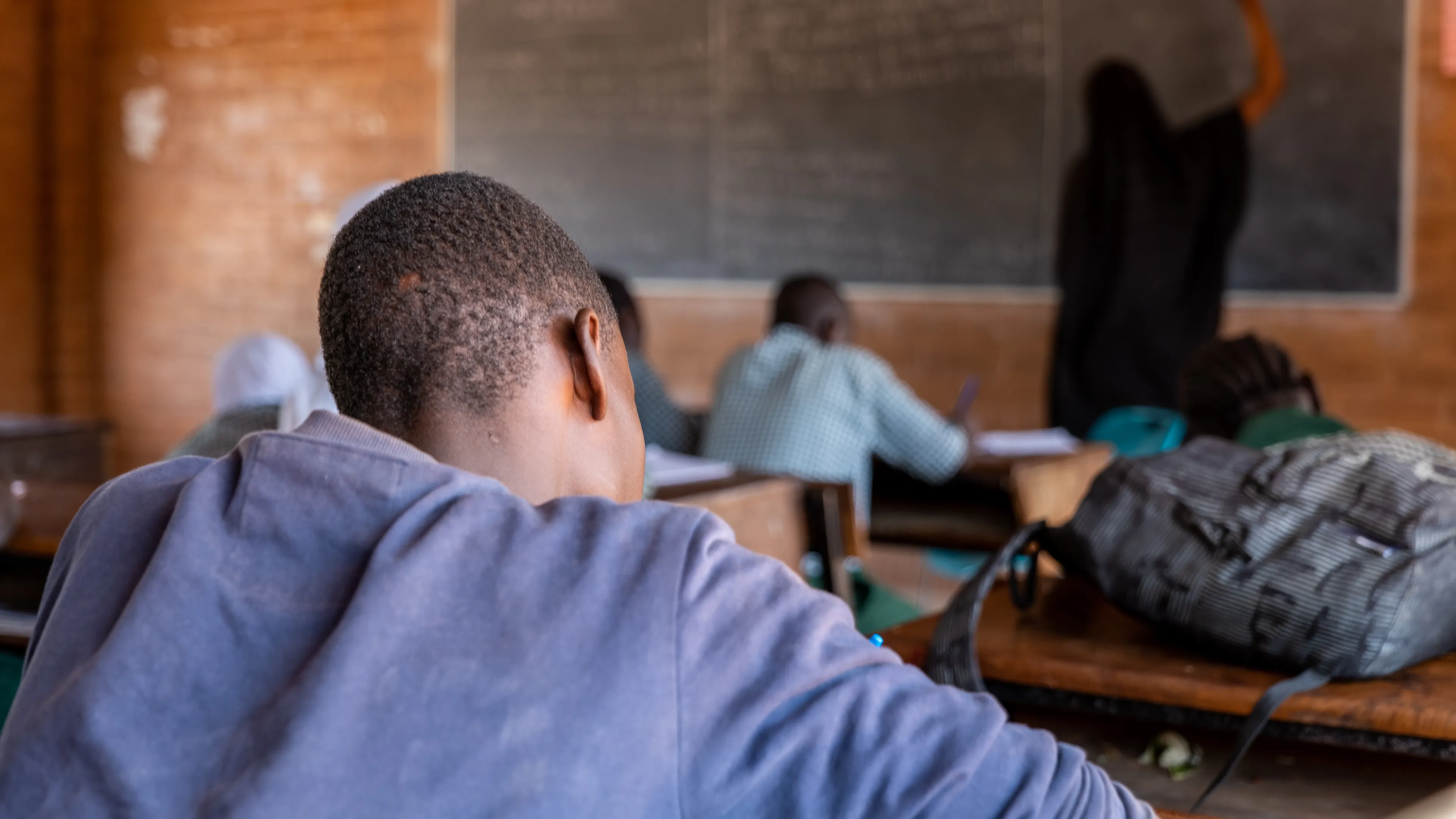 A classroom scene with students seated at desks, focusing on a blackboard at the front. A teacher, seen from behind, writes on the board. Backpacks and learning materials are visible on the desks.
