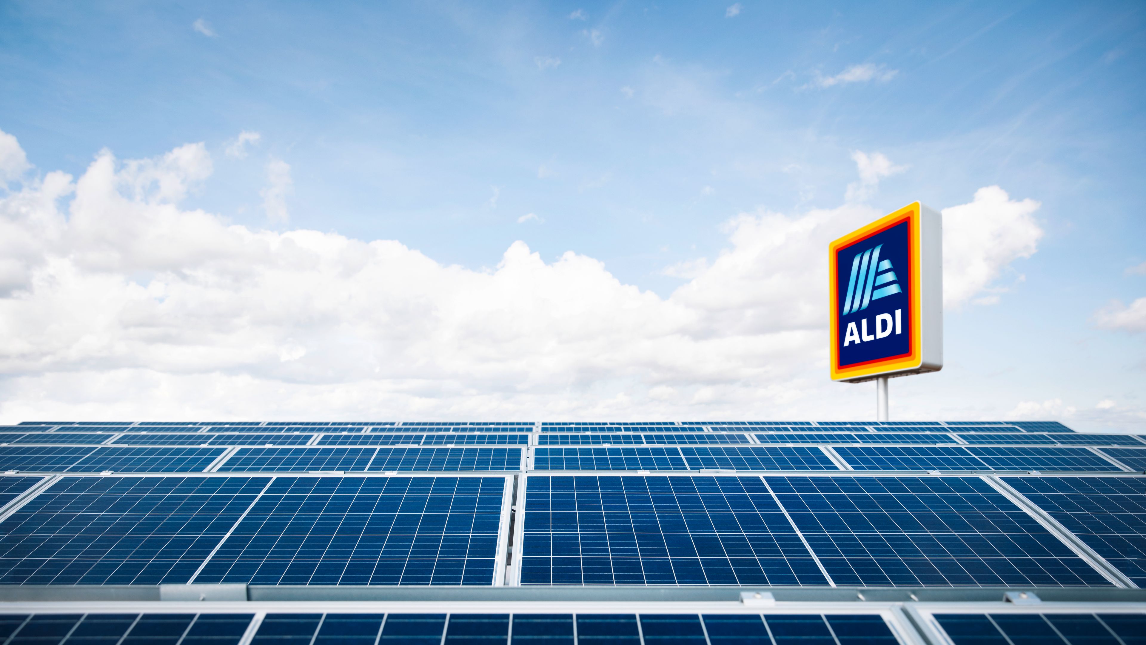 A solar panel array under a bright blue sky with scattered clouds. In the background, a sign with the Aldi logo is visible, indicating the location of the panels near an Aldi store.