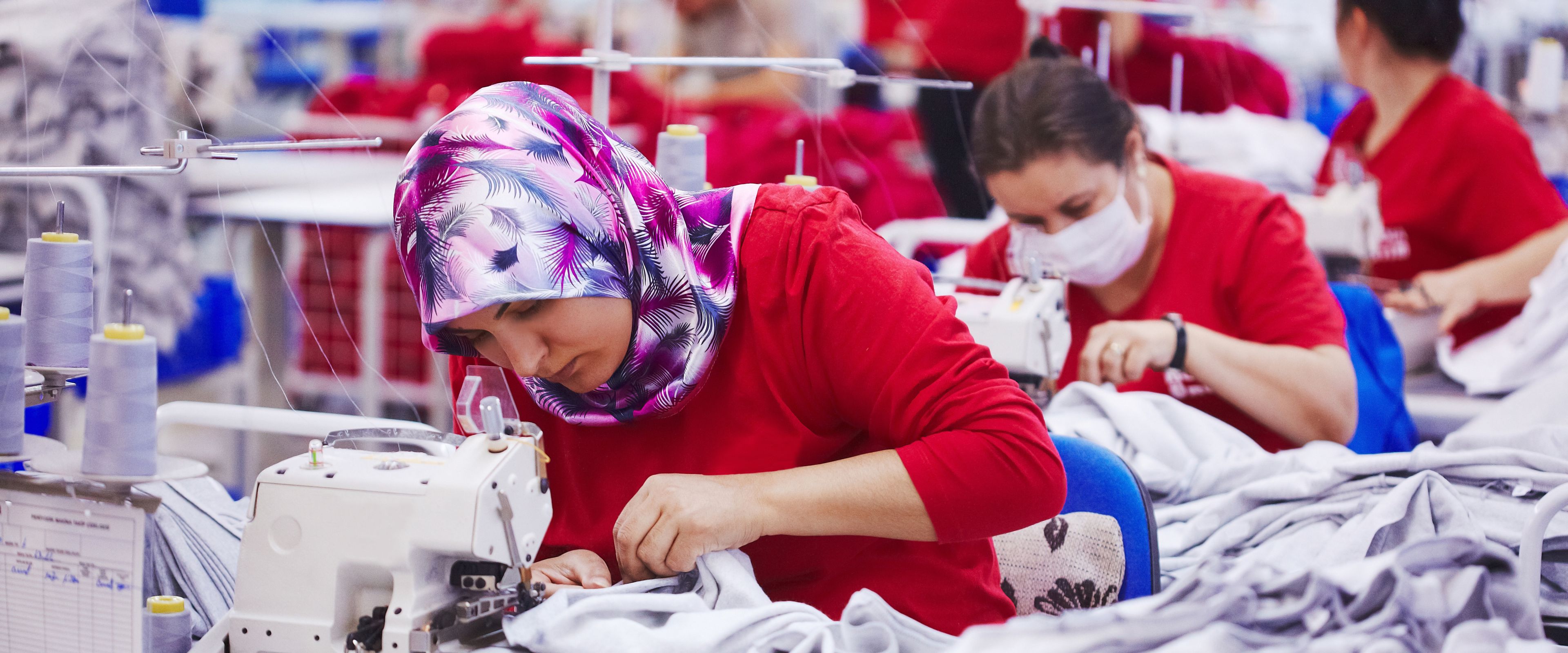 Several people are working in a textile factory on sewing machines. One woman in the foreground is wearing a colorful headscarf and focused on her work, while others are in the background, some wearing protective masks. The setting includes fabrics and industrial sewing equipment.