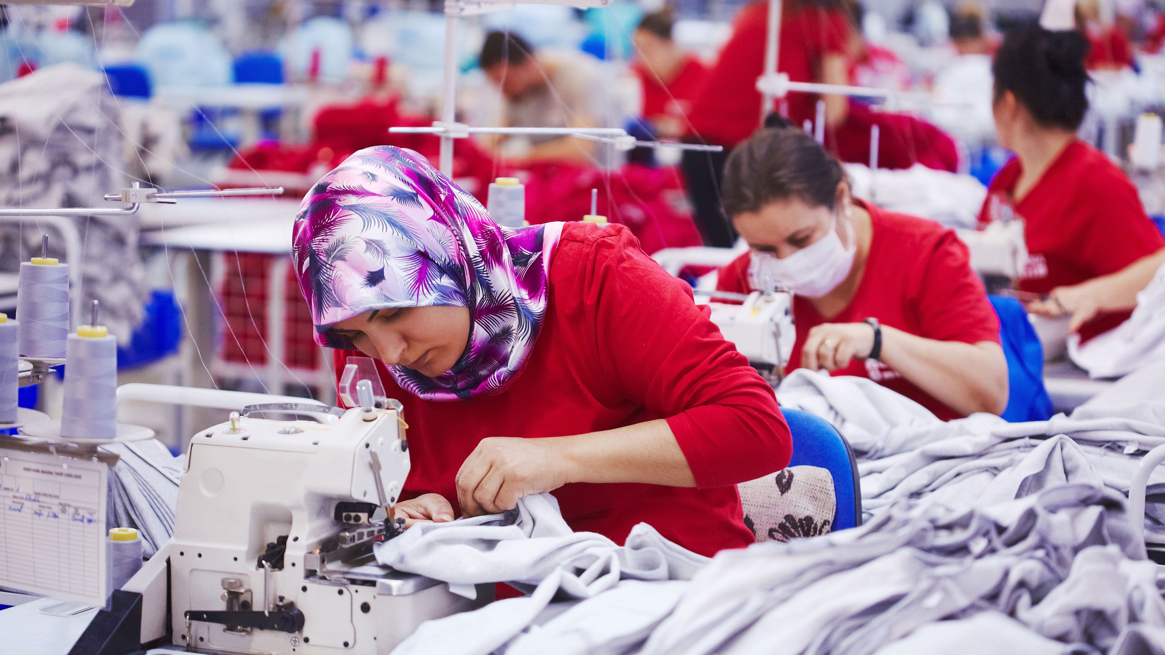 Several people are working in a textile factory on sewing machines. One woman in the foreground is wearing a colorful headscarf and focused on her work, while others are in the background, some wearing protective masks. The setting includes fabrics and industrial sewing equipment.