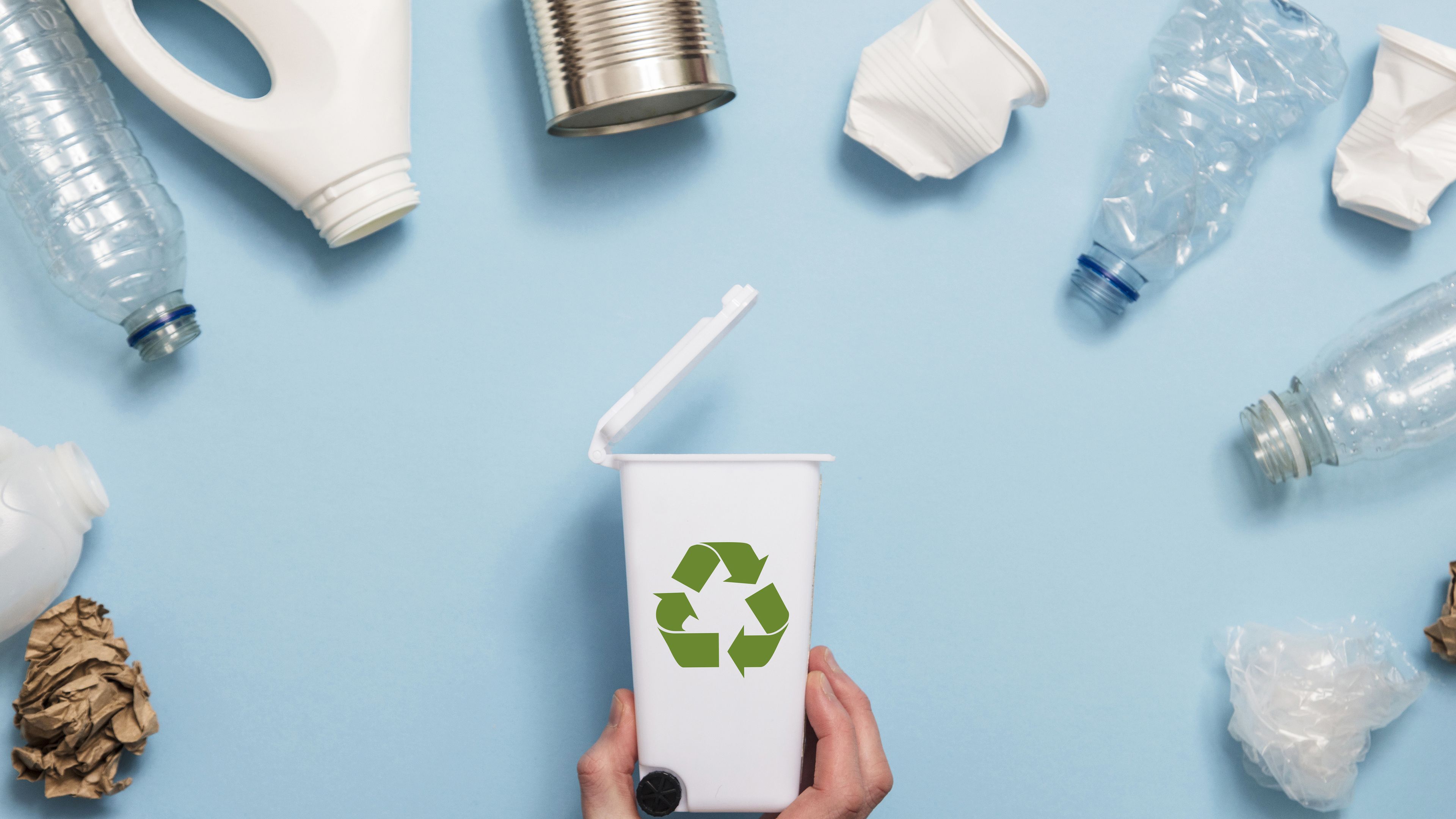 A hand holds a small white recycling bin with a green recycling symbol, surrounded by various recyclable materials like plastic bottles, cans, and paper on a light blue background. The bin’s lid is open, suggesting disposal of items. The image emphasizes recycling and environmental awareness.