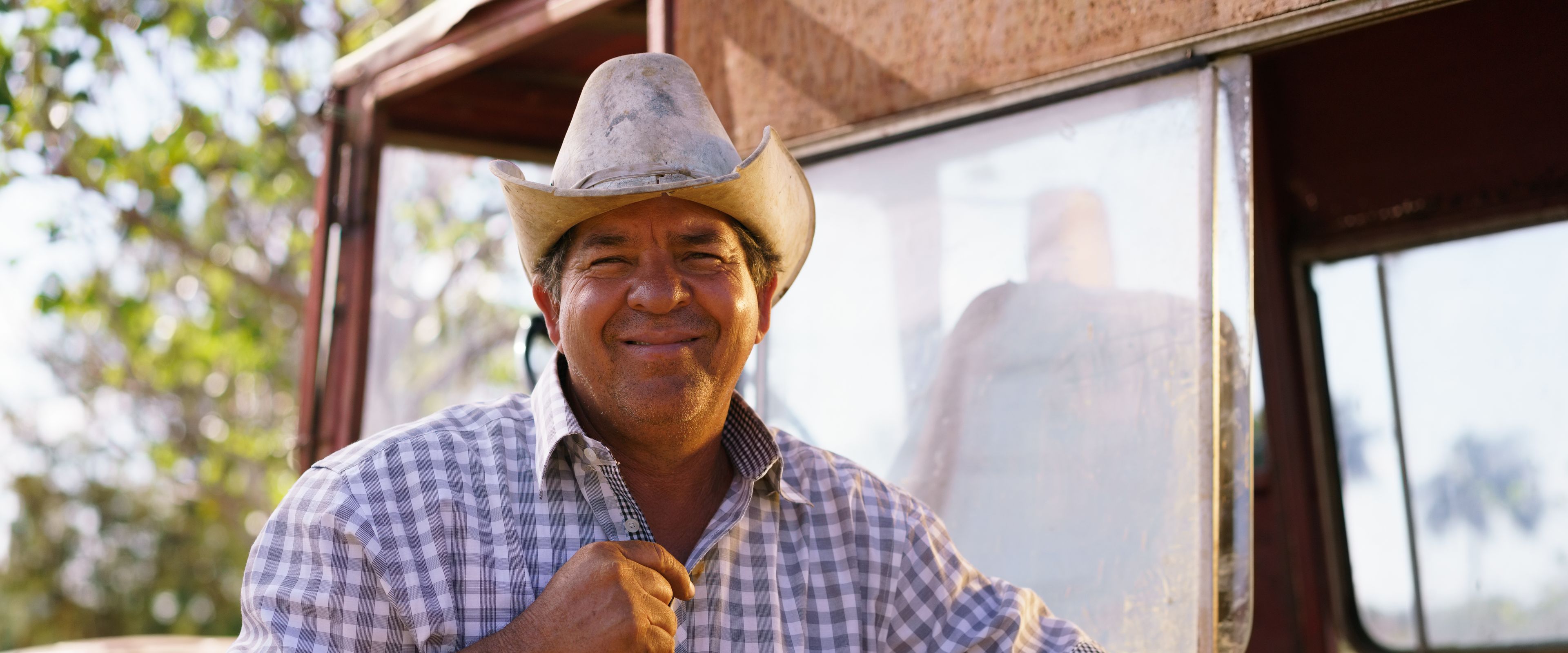 A man wearing a cowboy hat and a plaid shirt is smiling while leaning on a tool, possibly a shovel. He is standing next to a dusty vehicle in an outdoor setting with trees visible in the background. The scene suggests rural or agricultural surroundings.