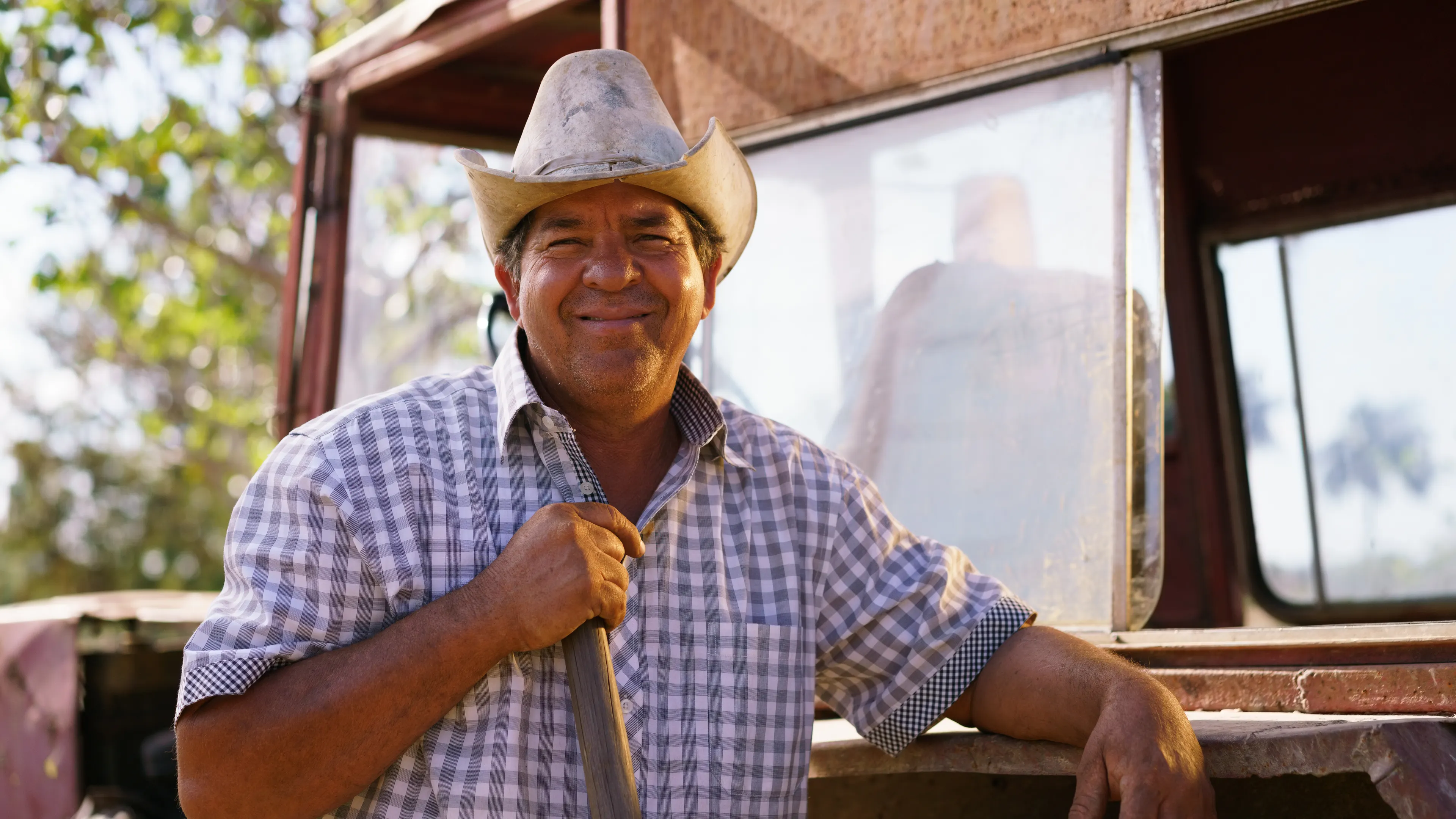 A man wearing a cowboy hat and a plaid shirt is smiling while leaning on a tool, possibly a shovel. He is standing next to a dusty vehicle in an outdoor setting with trees visible in the background. The scene suggests rural or agricultural surroundings.