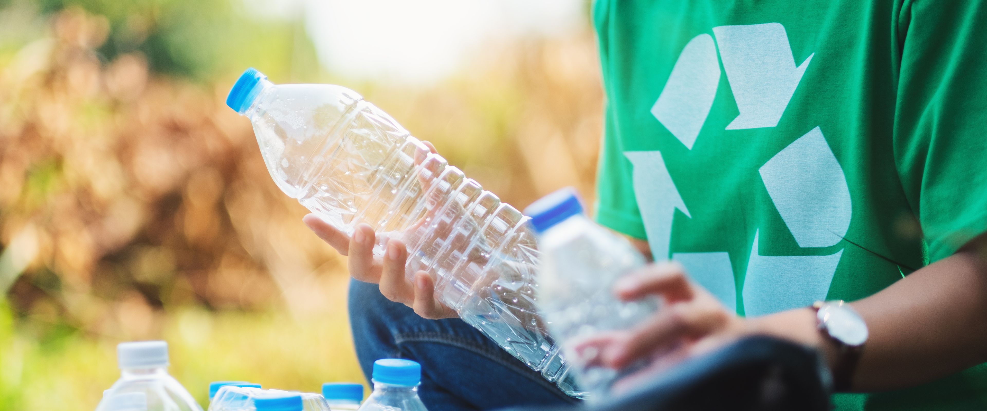 A person wearing a green shirt with a recycling symbol is holding a plastic bottle and sorting it into a container filled with other bottles. The scene takes place outdoors, surrounded by greenery. The activity appears focused on recycling and environmental care.