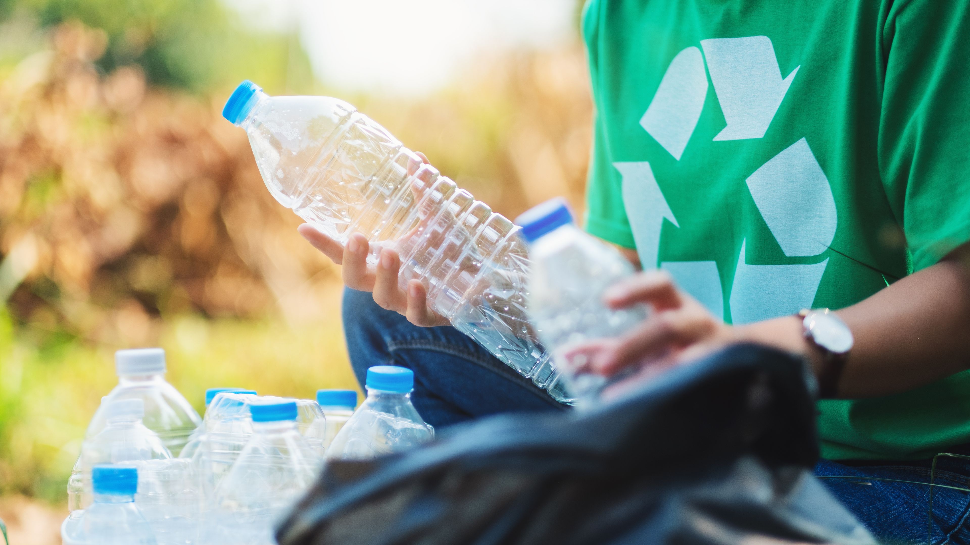 A person wearing a green shirt with a recycling symbol is holding a plastic bottle and sorting it into a container filled with other bottles. The scene takes place outdoors, surrounded by greenery. The activity appears focused on recycling and environmental care.