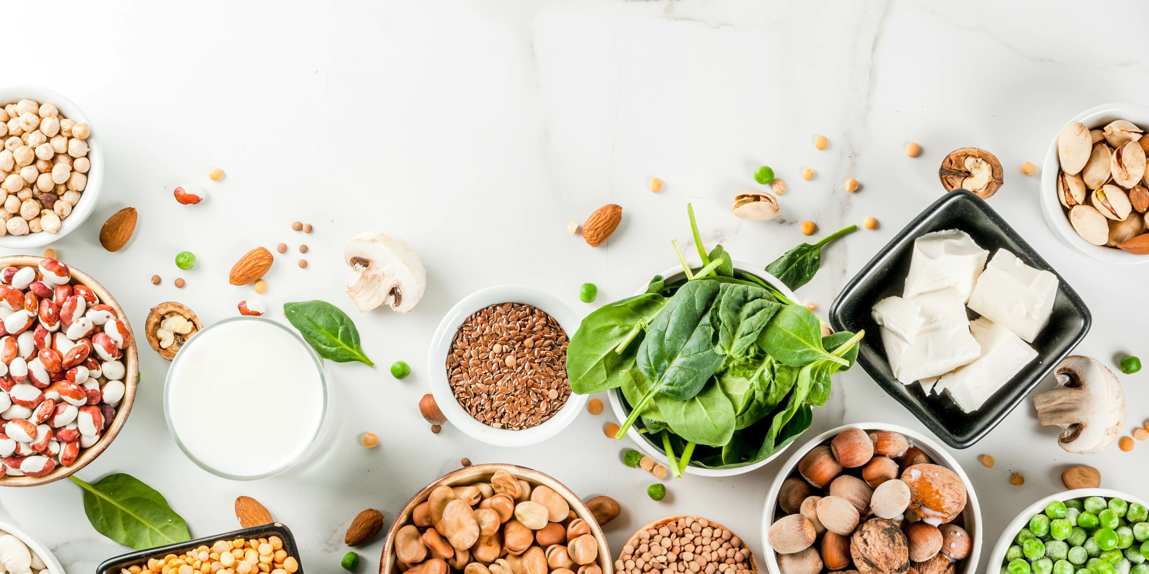 An assortment of plant-based protein sources is displayed on a white surface, including lentils, beans, spinach, tofu, nuts, and seeds in bowls. Additional scattered ingredients, like peas and almonds, enhance the arrangement. The overall scene promotes healthy, vegan nutrition.