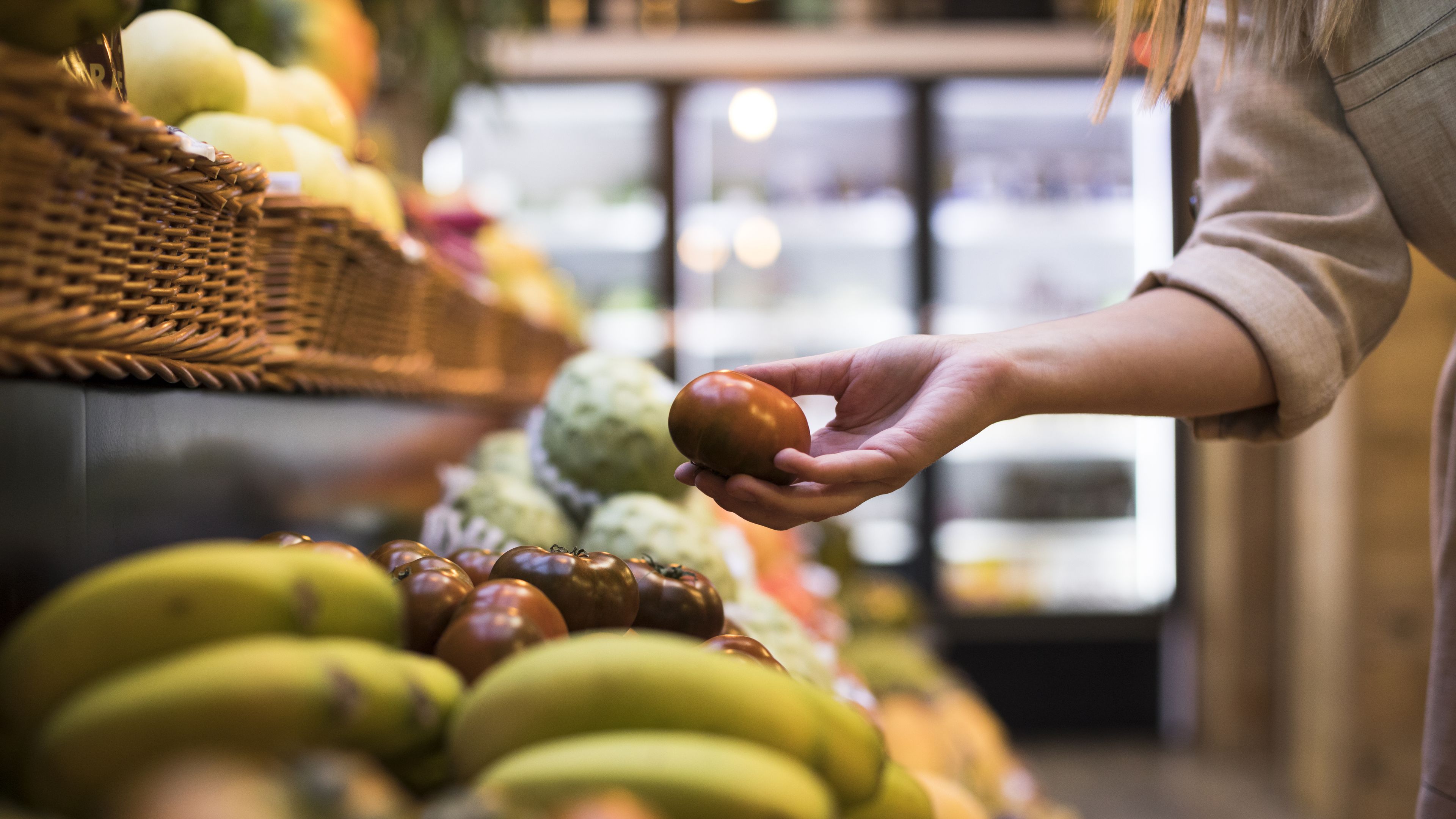 A person is picking up a small tomato from a display of fresh fruits and vegetables in a grocery store. The scene includes bananas, other produce, and baskets, with refrigerated sections visible in the background. The setting suggests a shopping activity.