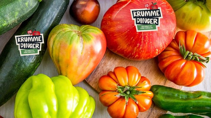 A variety of oddly shaped vegetables, including tomatoes, cucumbers, and peppers, are displayed on a light wooden surface. The text "KRUMME DINGER" appears prominently on a label, along with the tagline emphasizing taste over appearance. The scene promotes the appreciation of imperfect or misshapen produce.